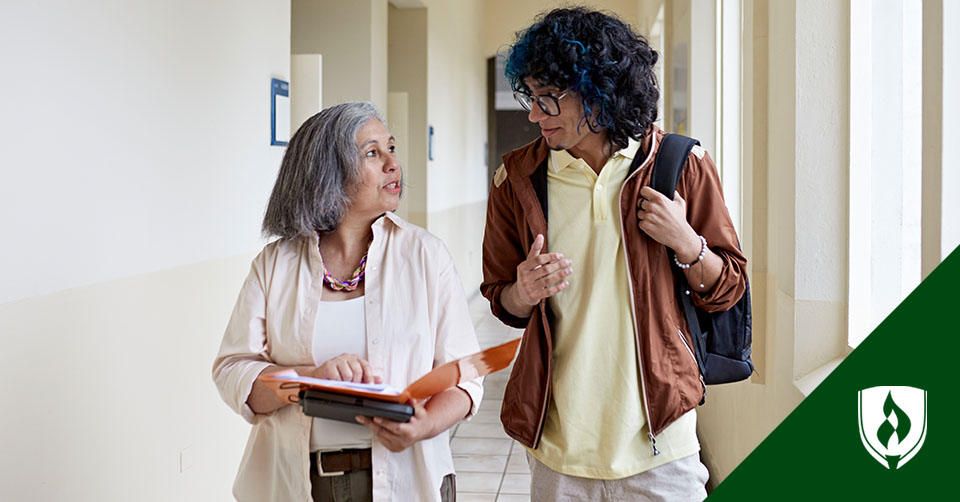 An older woman talks to a young man with blue in his hair as they walk down a hall.
