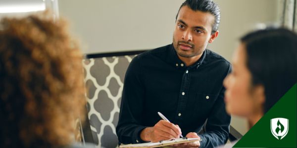 A caseworker sits with two clients, recording information on a clipboard