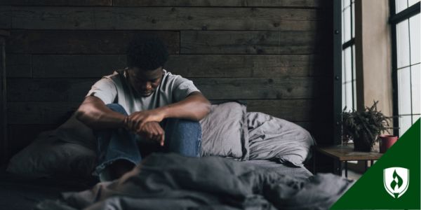 A man sits on his bed in a shadowed room looking lost